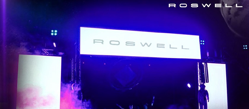 Escenario Roswell con pantallas, humo e iluminación violeta.